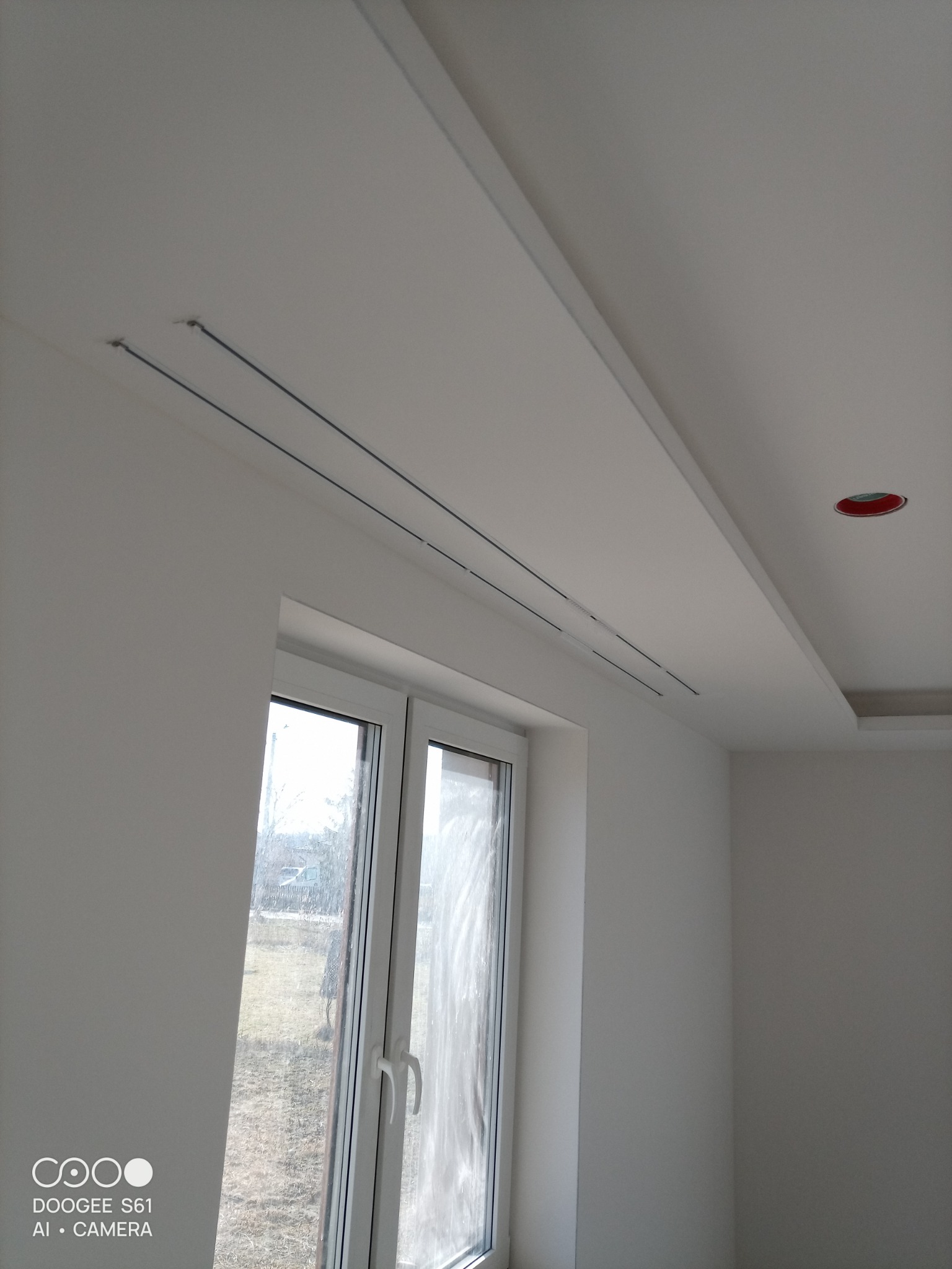 Białe wnętrze po remoncie: gładkie ściany, okno z widokiem na zewnątrz, sufit podwieszany z liniowymi nawiewnikami i punktowym oświetleniem. Minimalistyczny design.