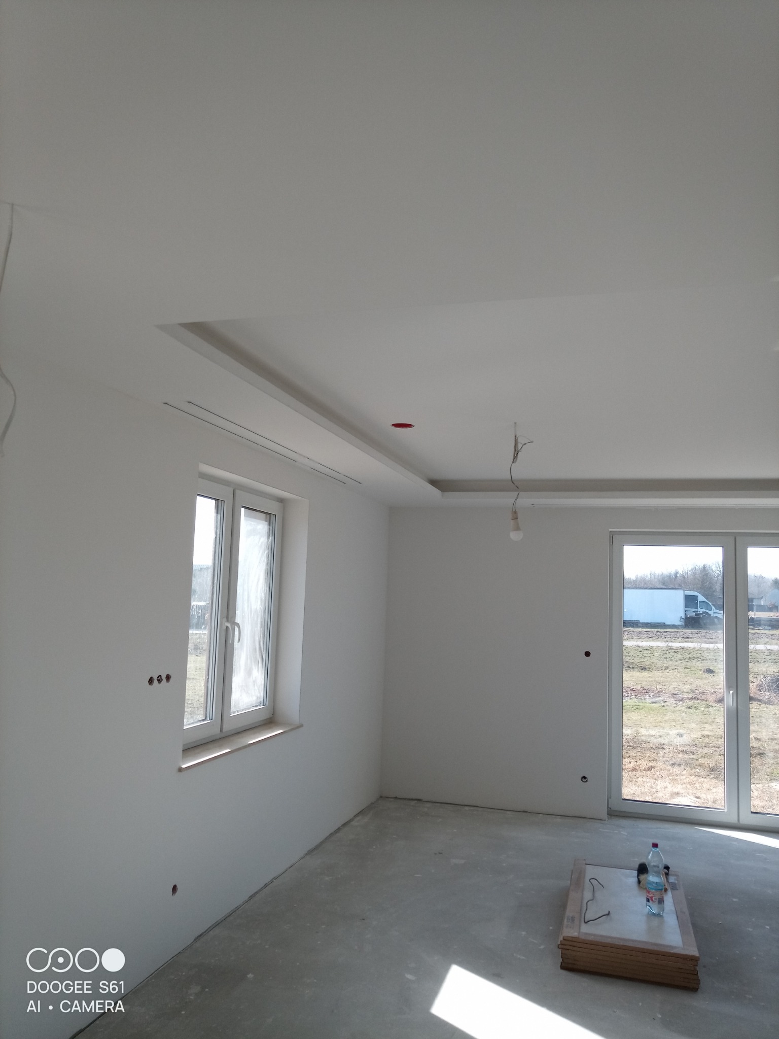 Wnętrze po gładziowaniu: białe ściany, sufit z podwieszanym elementem, okno i drzwi balkonowe. Na podłodze płyty i butelka wody. Widoczne ślady po gniazdkach.