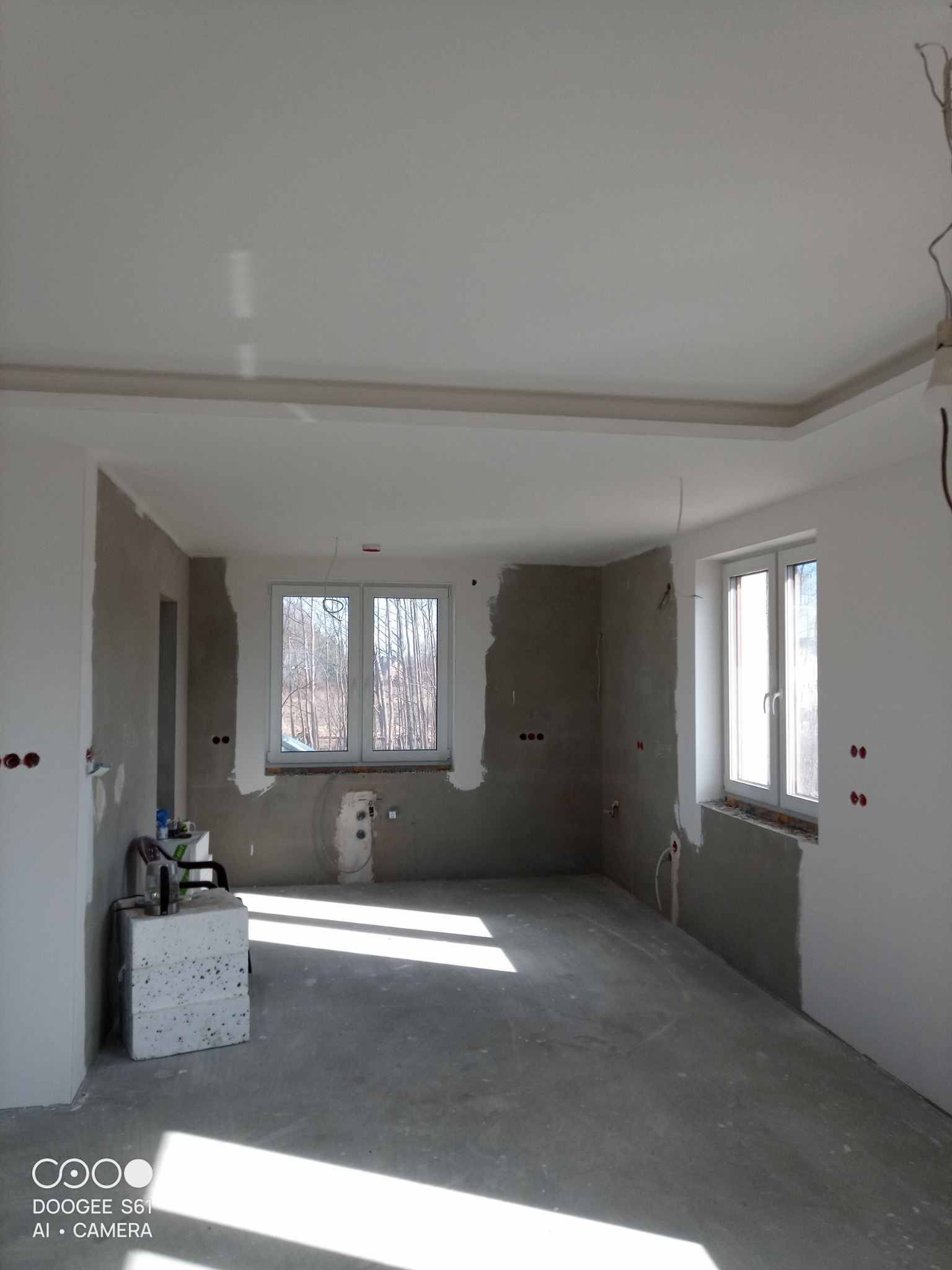 Wnętrze w trakcie remontu: surowe ściany przygotowane do malowania, widoczne instalacje elektryczne, okna z widokiem na drzewa, podłoga wylewka. Przygotowanie do wykończenia.