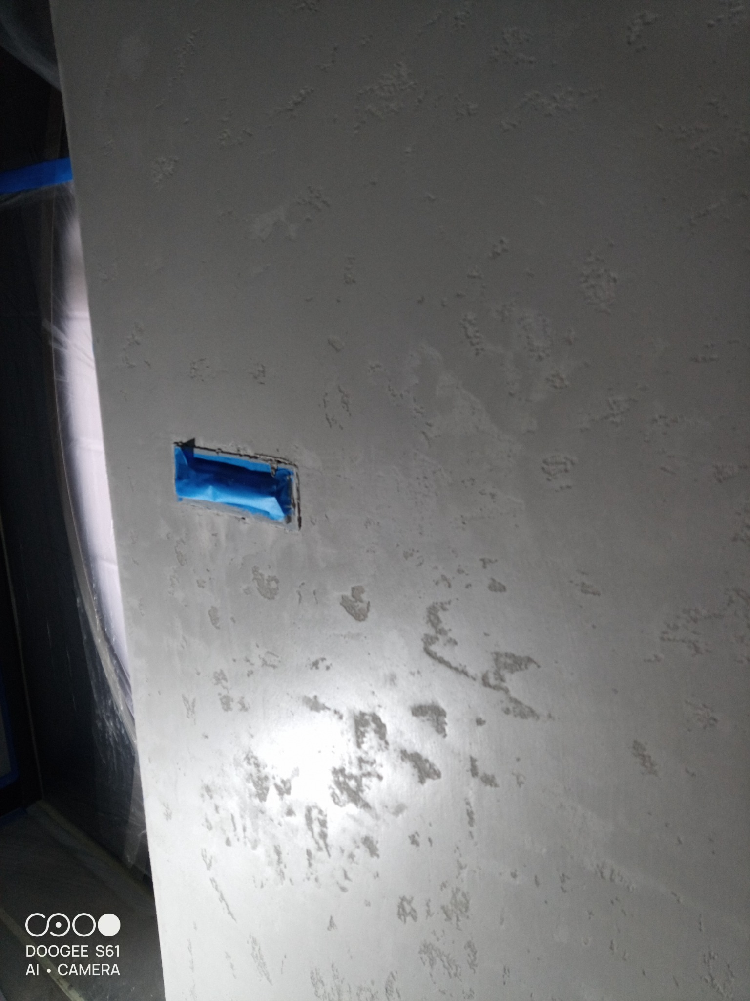 Ściana po szpachlowaniu z widocznym otworem na gniazdko zabezpieczonym taśmą malarską. Delikatna faktura na powierzchni. Przygotowanie do dalszych prac wykończeniowych.