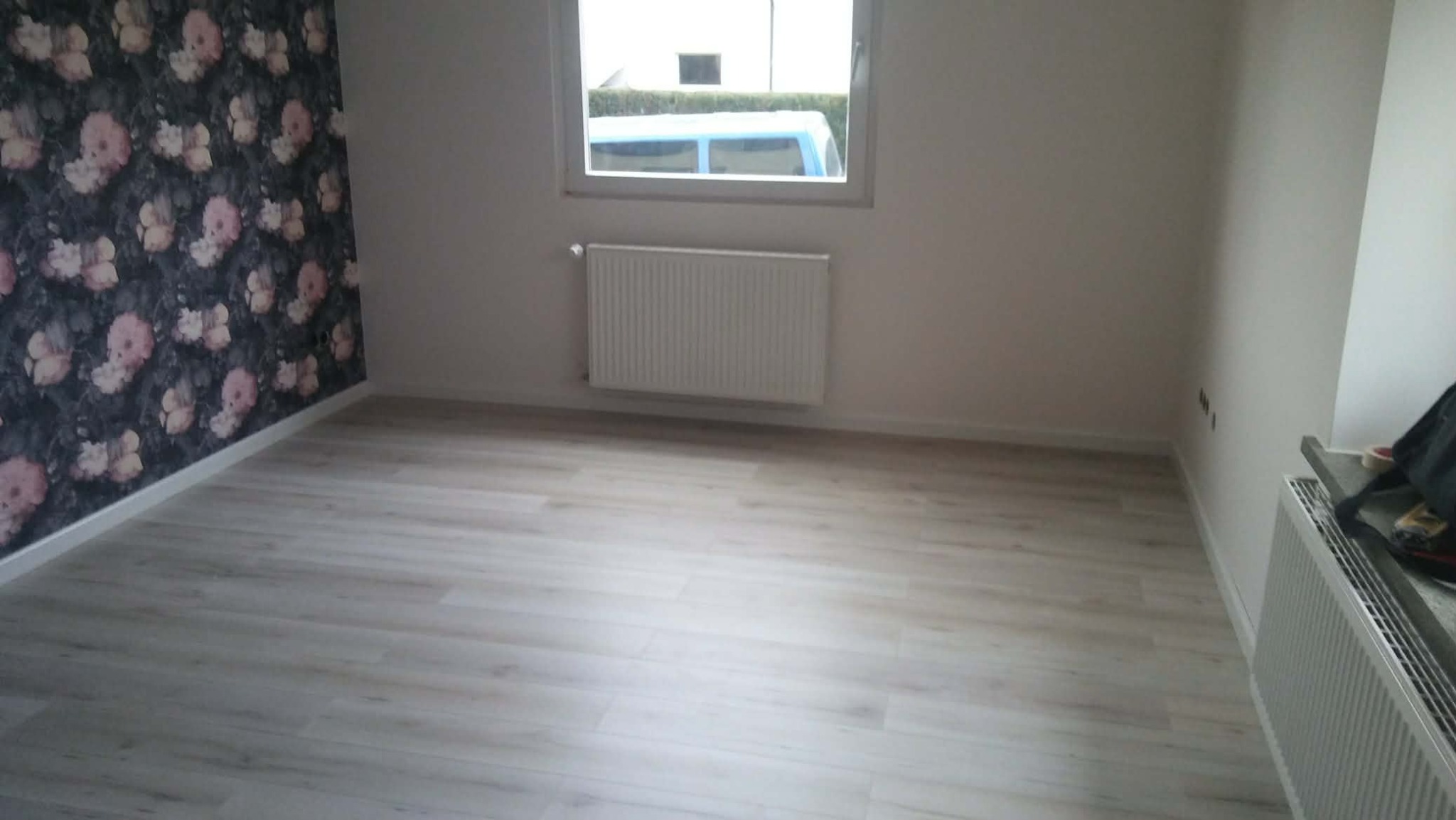 Jasne panele podłogowe w pokoju po remoncie. Ściana z tapetą w kwiaty, okno z grzejnikiem. Widoczne listwy przypodłogowe i gładkie ściany.