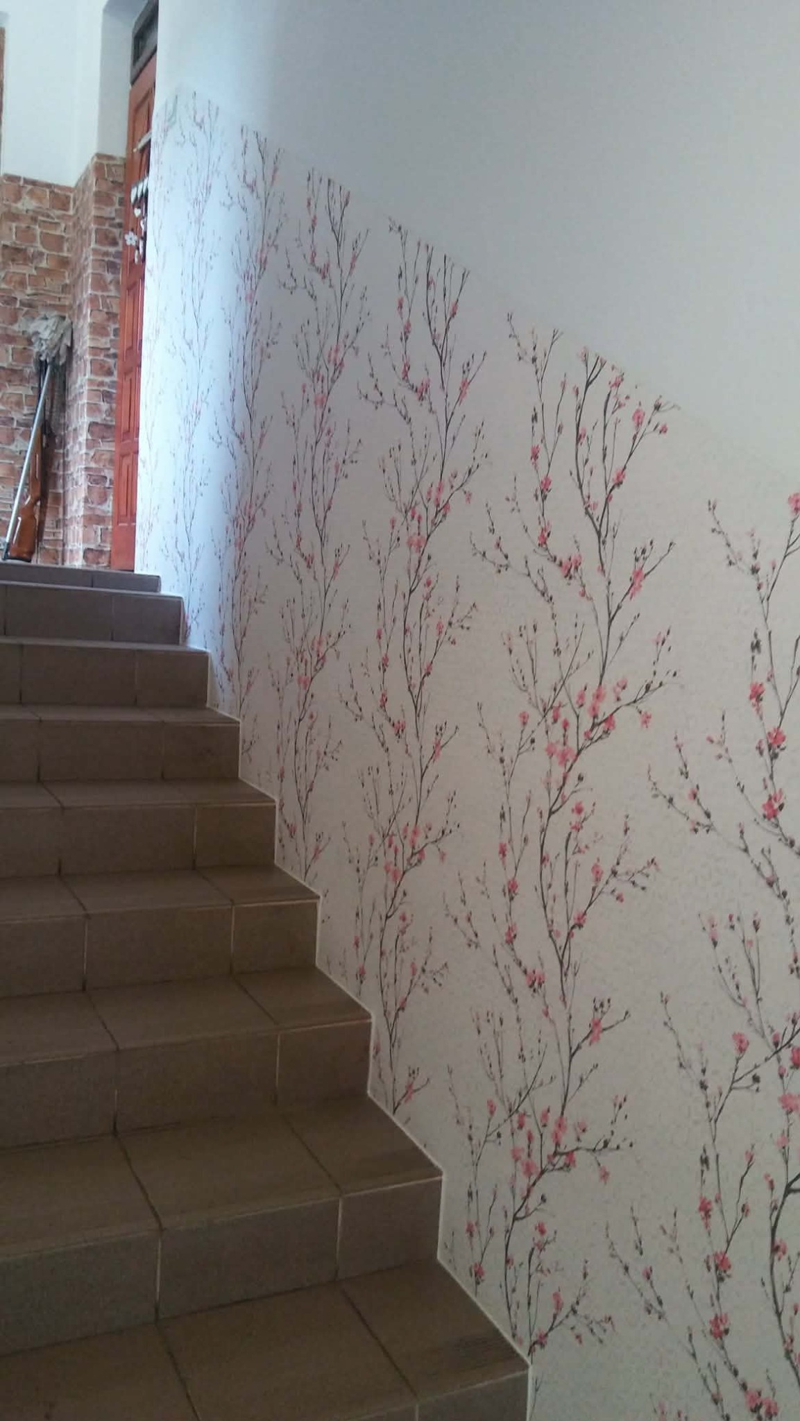 Klatka schodowa po malowaniu ścian z motywem kwitnącej wiśni. Brązowe płytki na schodach. Widoczny fragment ceglanej ściany i drzwi.