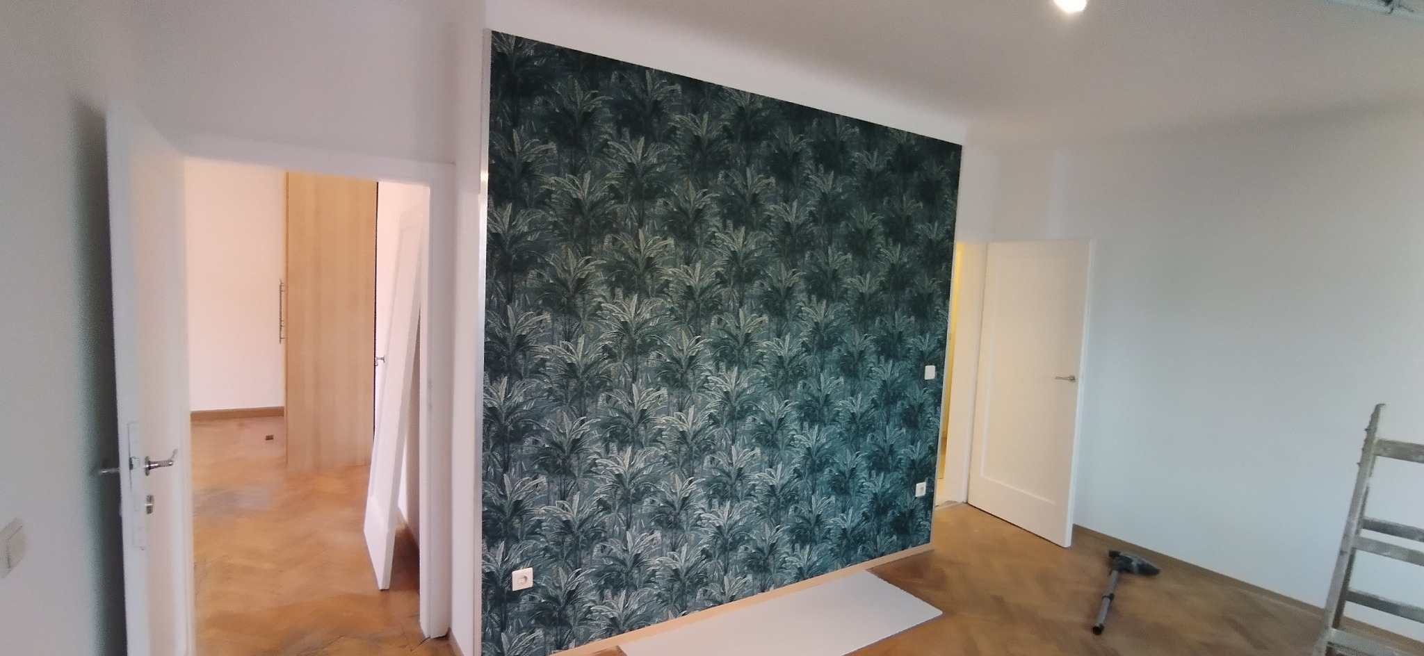 Pokój po remoncie: ściana z tapetą w roślinny wzór, drewniana podłoga, białe drzwi. Widoczna drabina i narzędzia, co sugeruje prace wykończeniowe.