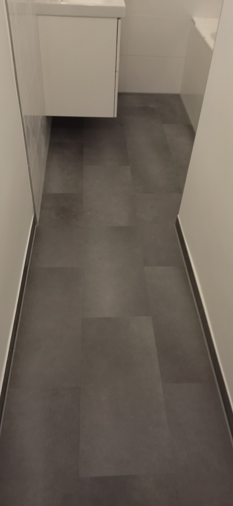 Ułożone, ciemnoszare panele podłogowe w wąskiej łazience z białą szafką pod umywalką. Widok z góry, prosto, z delikatnym oświetleniem.