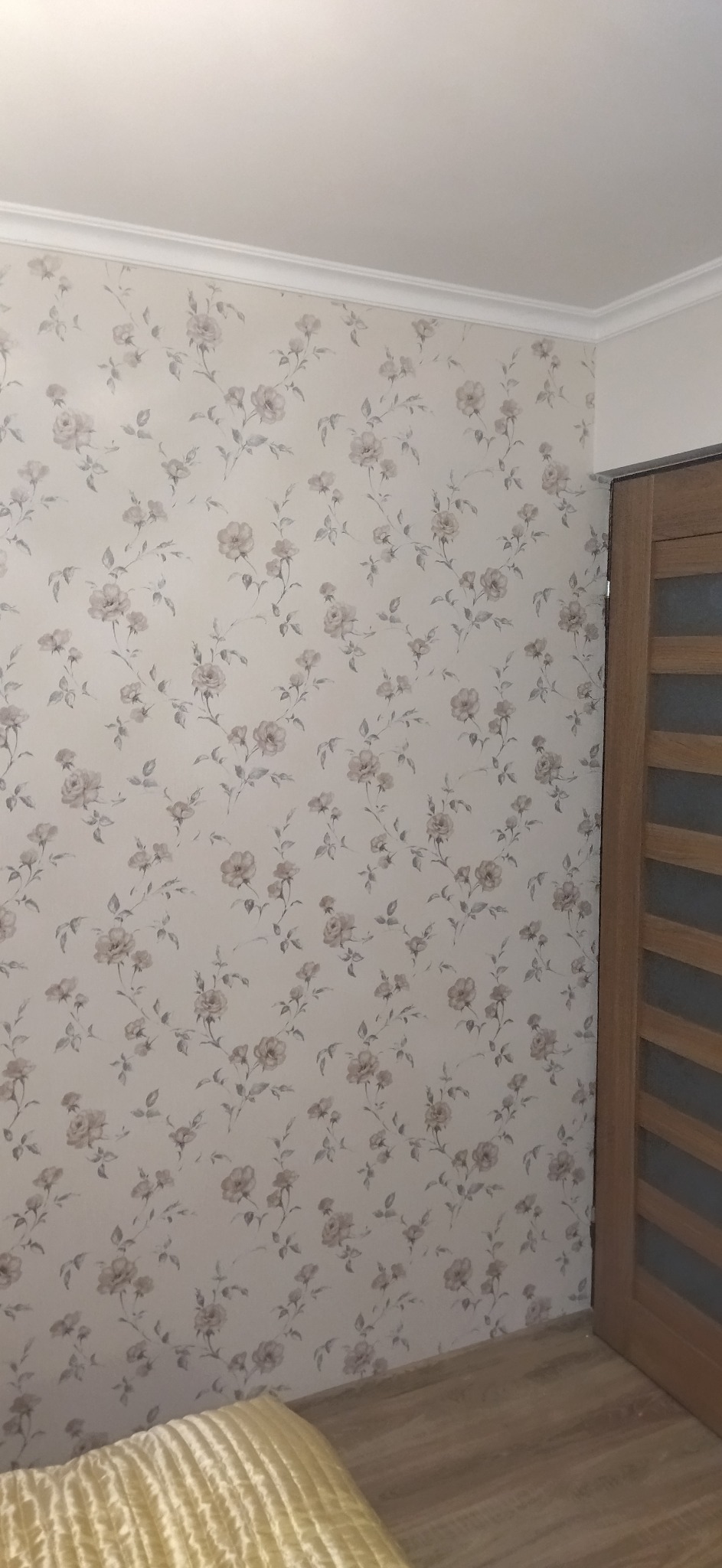 Ściana z tapetą w kwiaty i drewniane drzwi. Widoczny fragment sufitu z listwą dekoracyjną. U dołu kadru fragment łóżka z żółtą narzutą.