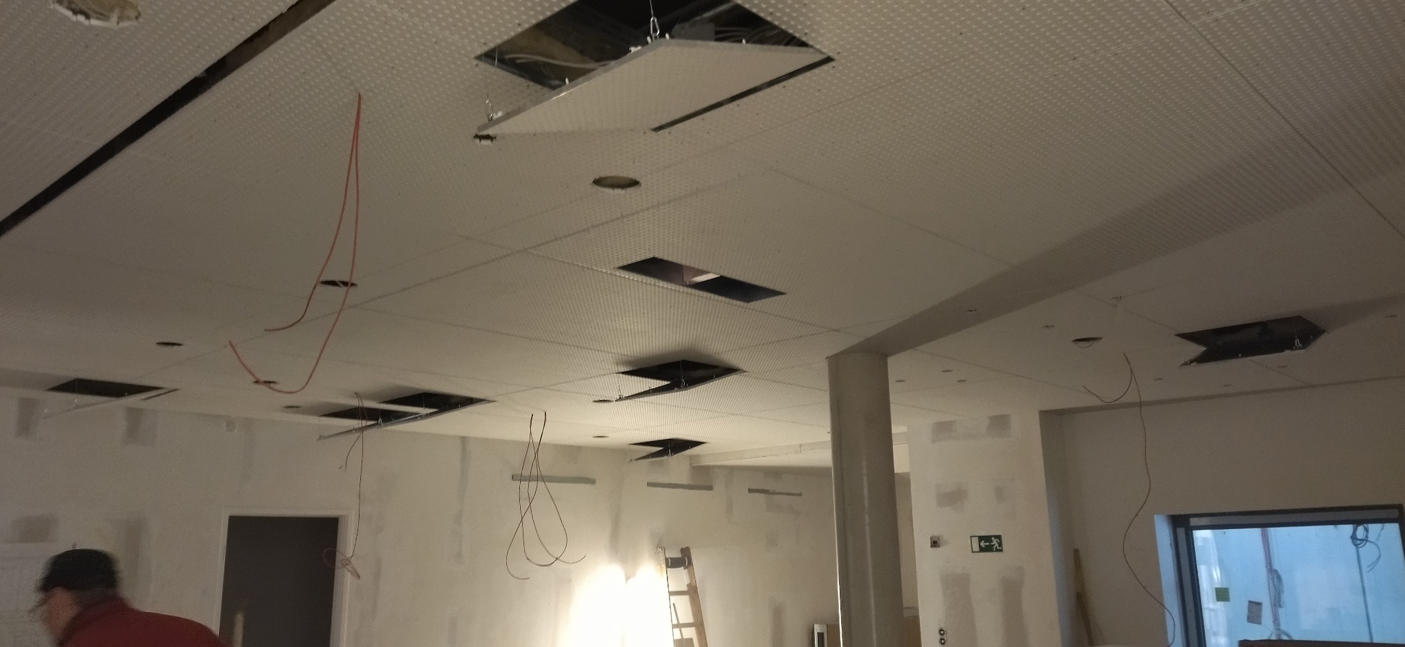 Sufit podwieszany z odkrytymi fragmentami, widoczne kable i drabina. W tle ściany w trakcie szpachlowania. Prace wykończeniowe w pomieszczeniu.