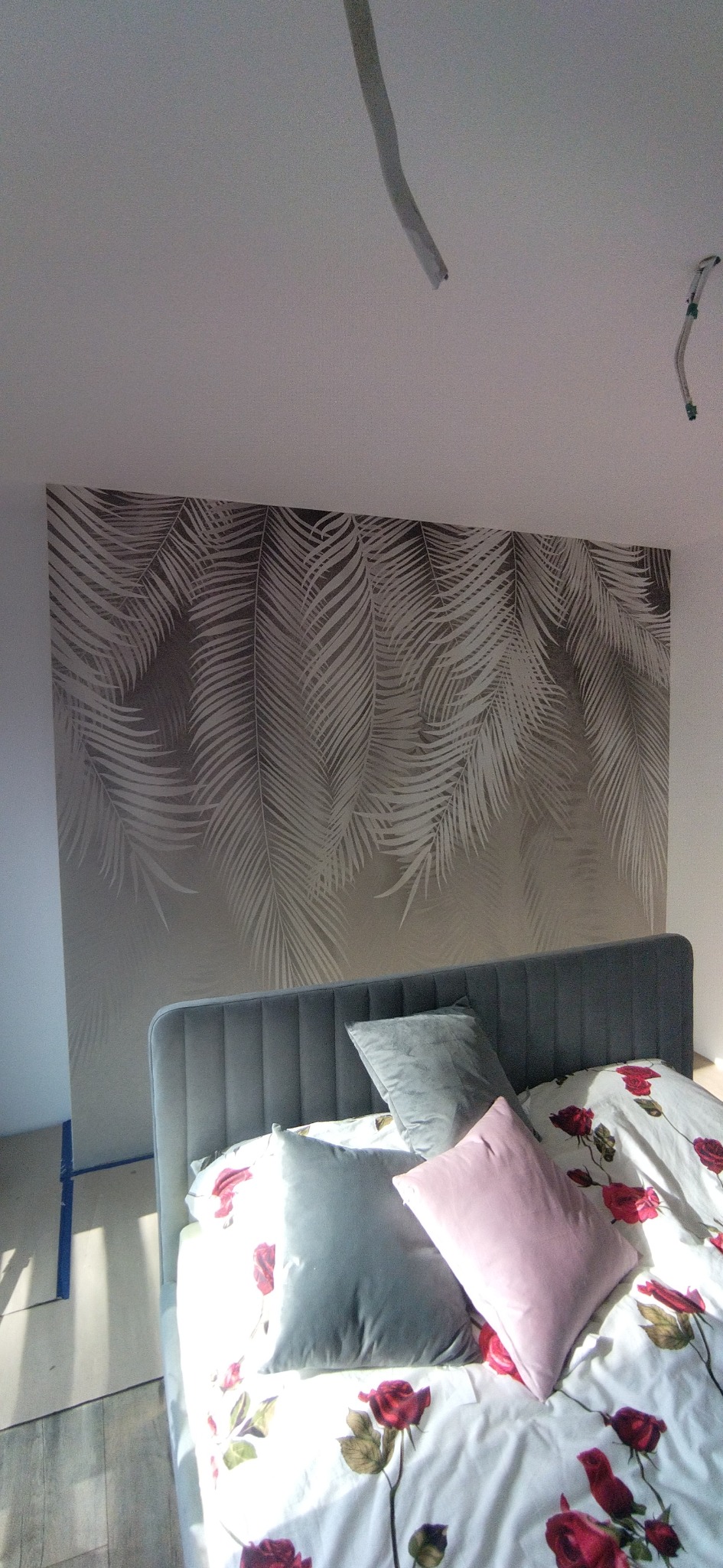 Sypialnia z łóżkiem i tapetą w liście palmowe. Widoczne niedokończone prace wykończeniowe, rury na suficie. Ujęcie z dołu, jasne światło.