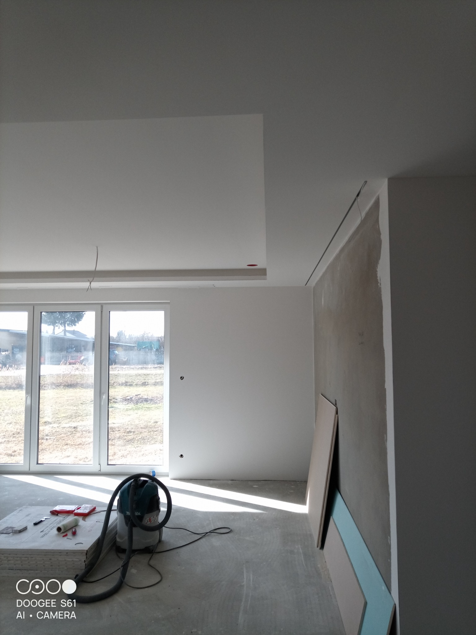 Wnętrze w trakcie prac wykończeniowych: surowa ściana, płyty gipsowe oparte o ścianę, odkurzacz budowlany i narzędzia na podłodze. Widoczne okno z widokiem na zewnątrz.