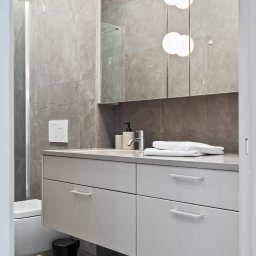 Michał Kotyński - Nowoczesna łazienka z szarymi płytkami imitującymi marmur, białą szafką pod umywalką i podwieszaną toaletą. Widoczny czarny kosz na śmieci i oświetlenie punktowe.