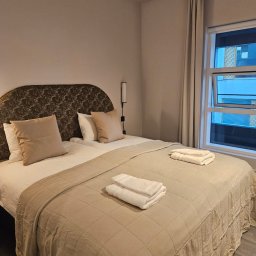 Michał Kotyński - Czysty pokój hotelowy z dwoma łóżkami, beżową narzutą i białymi ręcznikami. Widok na okno z zasłonami i fragment budynku za nim. Minimalistyczny wystrój.