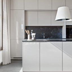 Michał Kotyński - Biała, nowoczesna kuchnia z czarnym blatem i marmurowym panelem. Minimalistyczny design, jasne światło, drewniane przybory kuchenne na blacie. Elegancki wystrój wnętrza.
