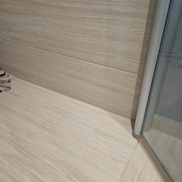 Michał Kotyński - Jasna, drewnopodobna podłoga i ściana w łazience, z fragmentem kabiny prysznicowej. Widoczne detale wykończenia i połączenie materiałów.