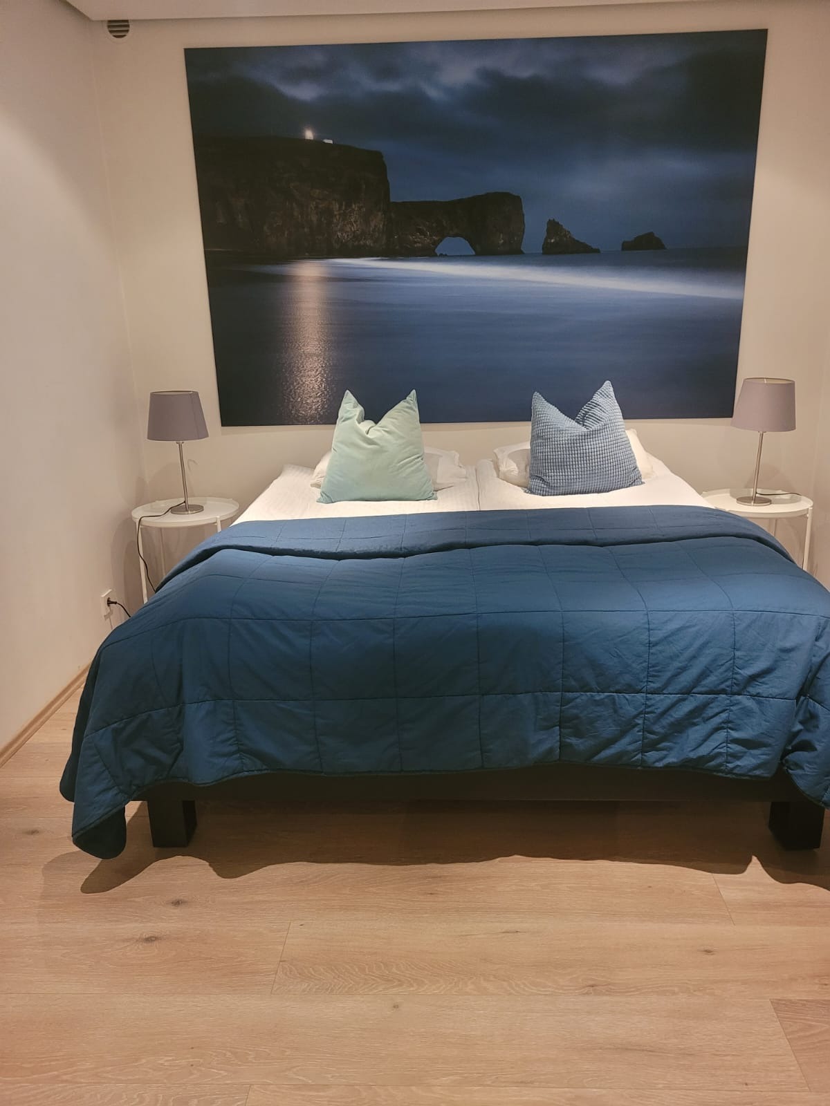 Dwuosobowe łóżko z niebieską narzutą i poduszkami, nocne stoliki z lampkami. Nad łóżkiem duży obraz z motywem morskim. Parkiet na podłodze. Czyste i schludne wnętrze.