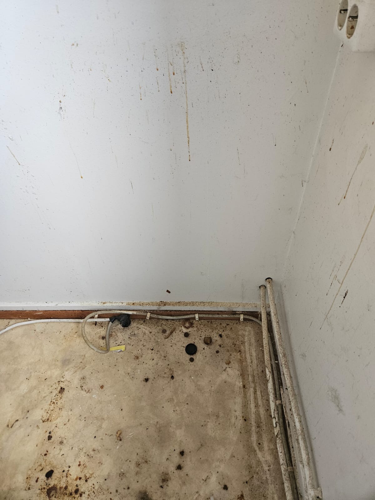 Zabrudzone pomieszczenie w trakcie remontu: ślady farby na ścianach, resztki gruzu na podłodze, widoczne rury i kable. Stan przed sprzątaniem poremontowym.