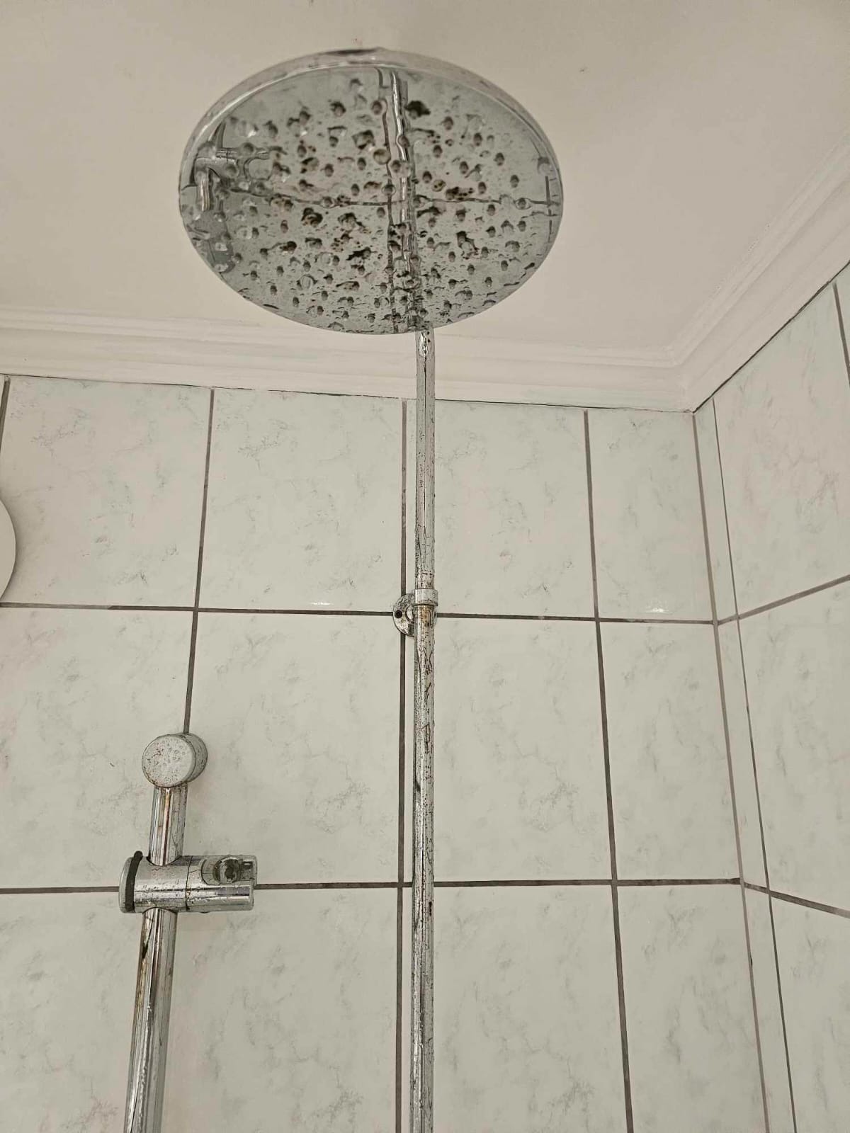Zardzewiała, chromowana deszczownica z widocznymi śladami kamienia i zaciekami, zamontowana na tle białych, kwadratowych płytek w łazience. Widoczne osady i brak połysku.