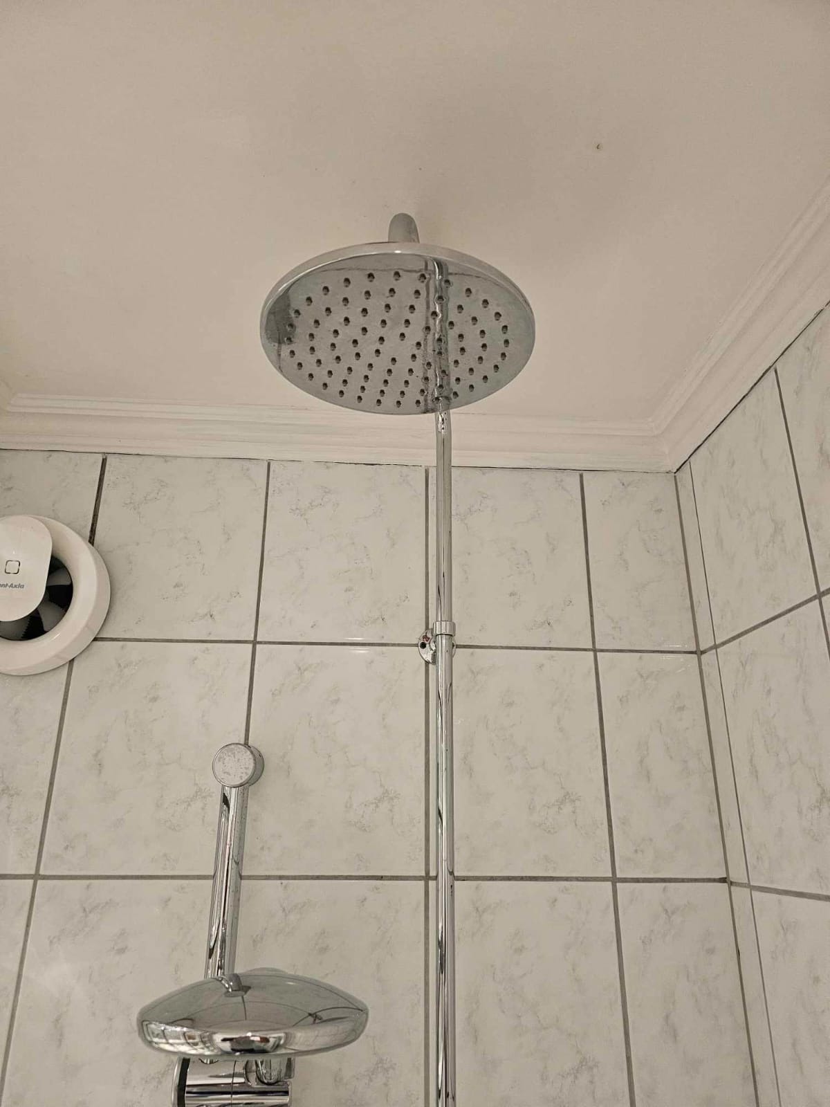 Biała łazienka z chromowaną armaturą prysznicową i wentylatorem. Widoczne płytki ścienne z delikatnym wzorem. Minimalistyczny design.