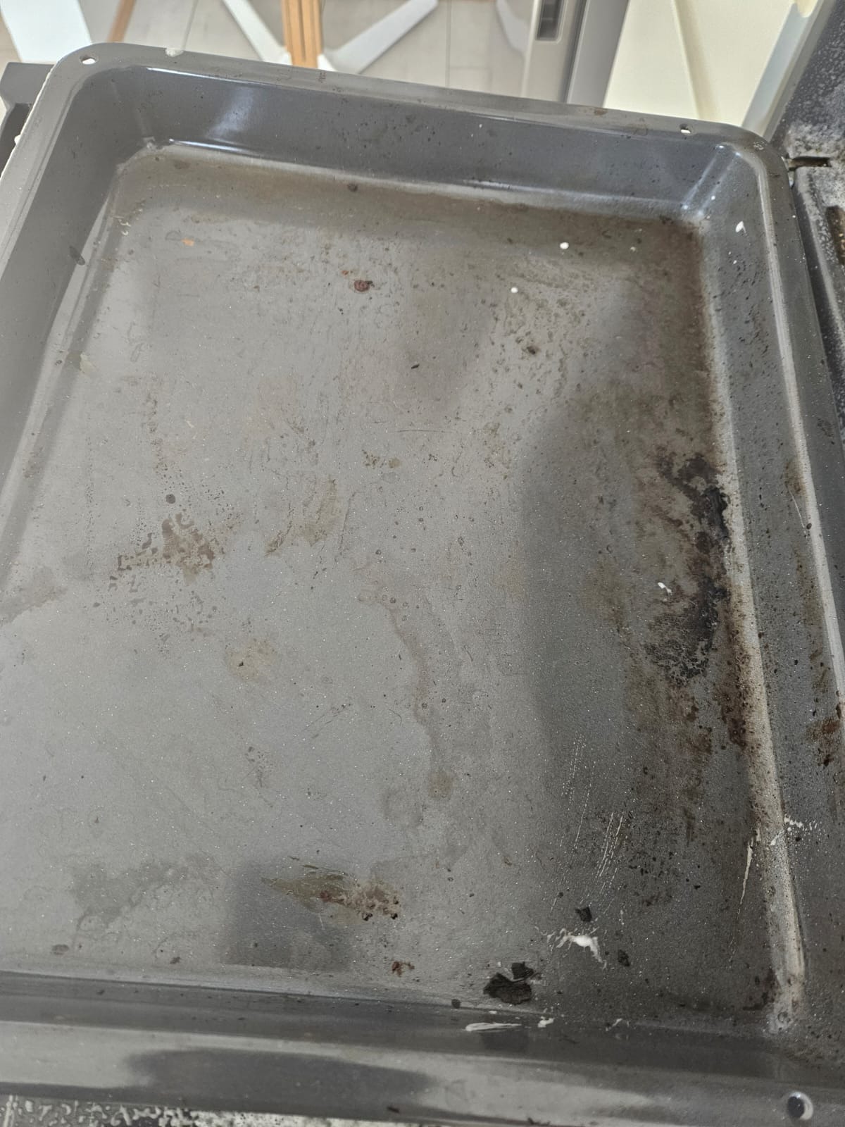 Zabrudzona blacha piekarnika z przypalonymi resztkami jedzenia, wymagająca dokładnego czyszczenia. Widoczne plamy i osady na szarej powierzchni blachy.