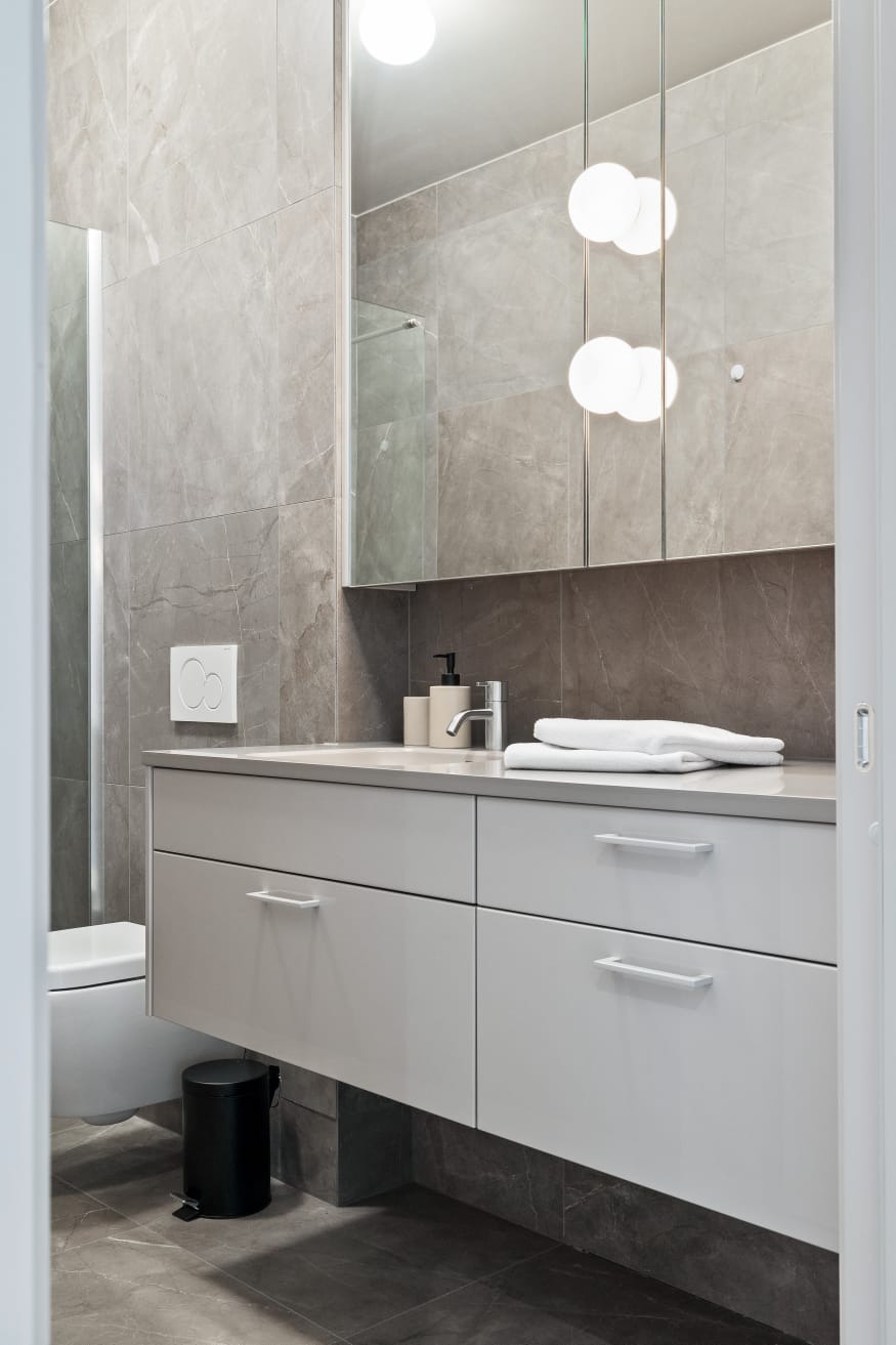 Nowoczesna łazienka z szarymi płytkami imitującymi marmur, białą szafką pod umywalką i podwieszaną toaletą. Widoczny czarny kosz na śmieci i oświetlenie punktowe.