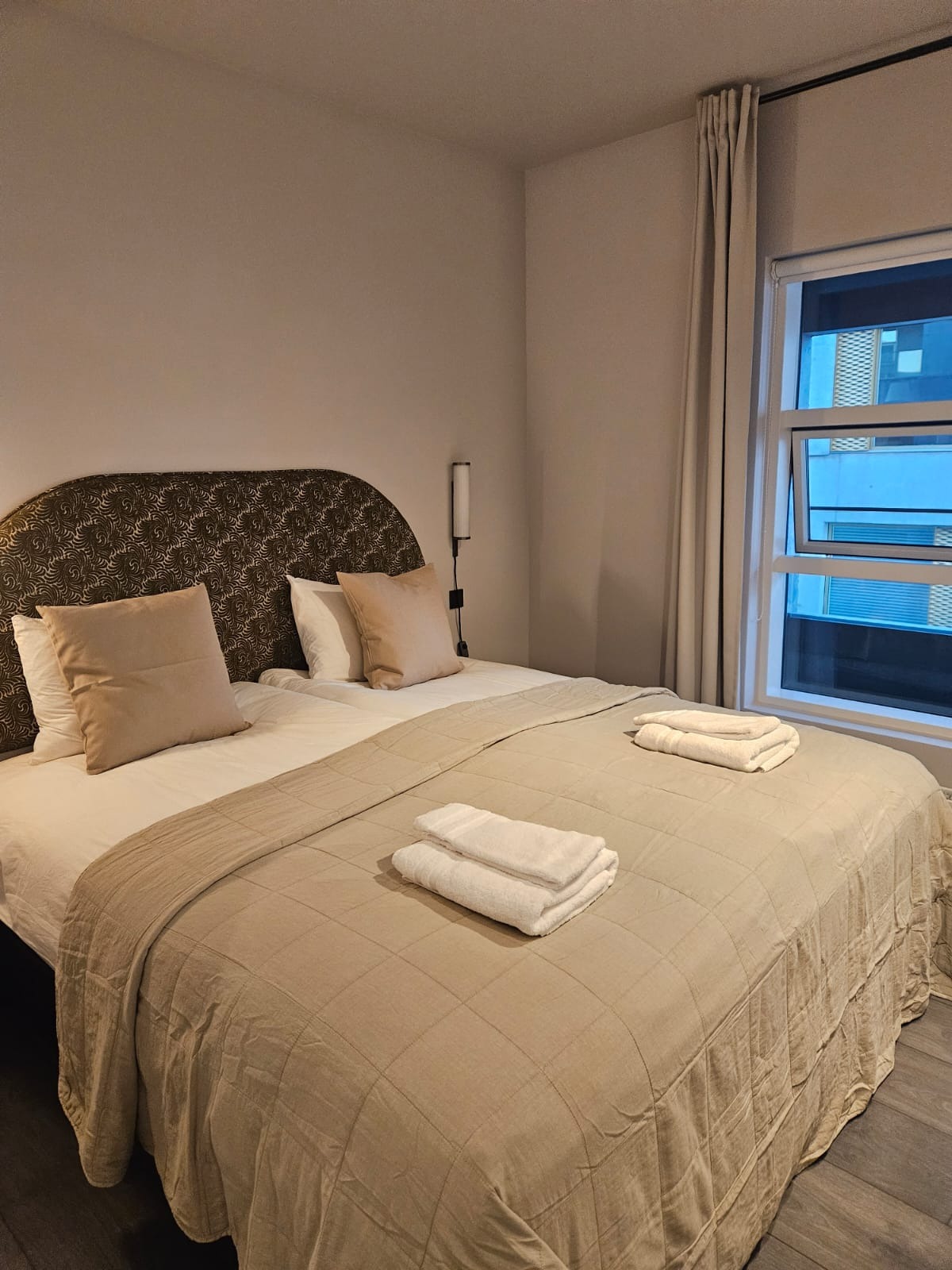 Czysty pokój hotelowy z dwoma łóżkami, beżową narzutą i białymi ręcznikami. Widok na okno z zasłonami i fragment budynku za nim. Minimalistyczny wystrój.