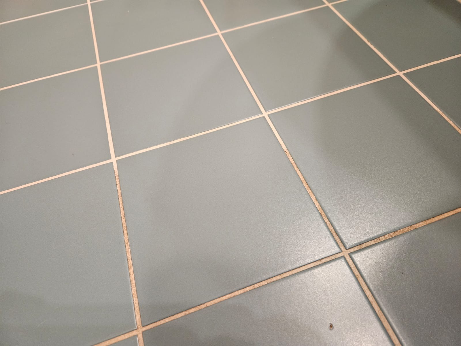 Błękitne, kwadratowe płytki podłogowe z jasnymi fugami, widoczne z bliska pod kątem. Fugi miejscami zabrudzone, co sugeruje potrzebę czyszczenia.
