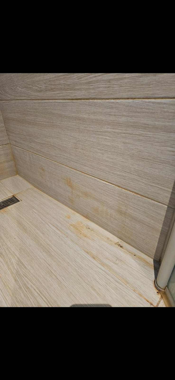 Zabrudzona kabina prysznicowa z jasnymi płytkami imitującymi drewno. Widoczne zacieki i osady na fugach oraz płytkach podłogowych i ściennych. Narożnik kabiny.