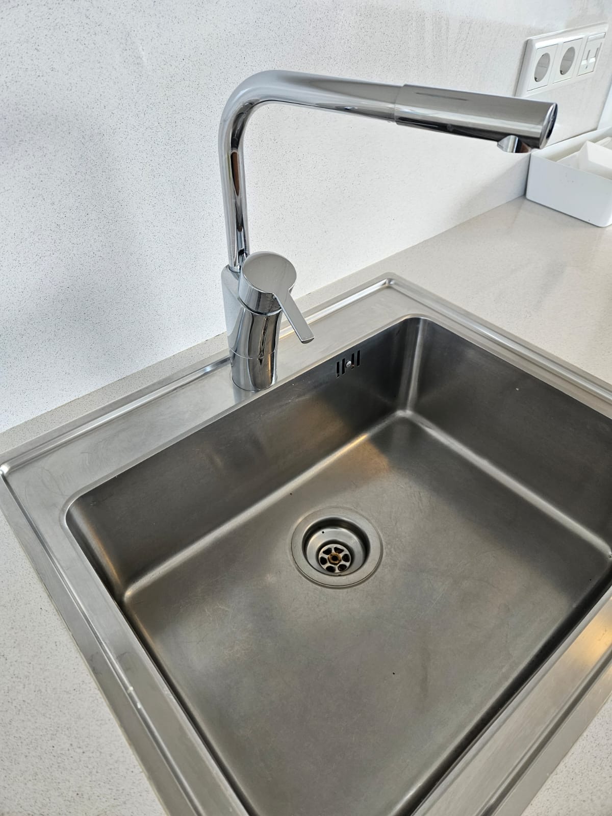 Błyszczący, chromowany kran nad czystym, metalowym zlewem w kuchni. Widoczne drobne ślady użytkowania, podkreślające autentyczność. Minimalistyczne tło.