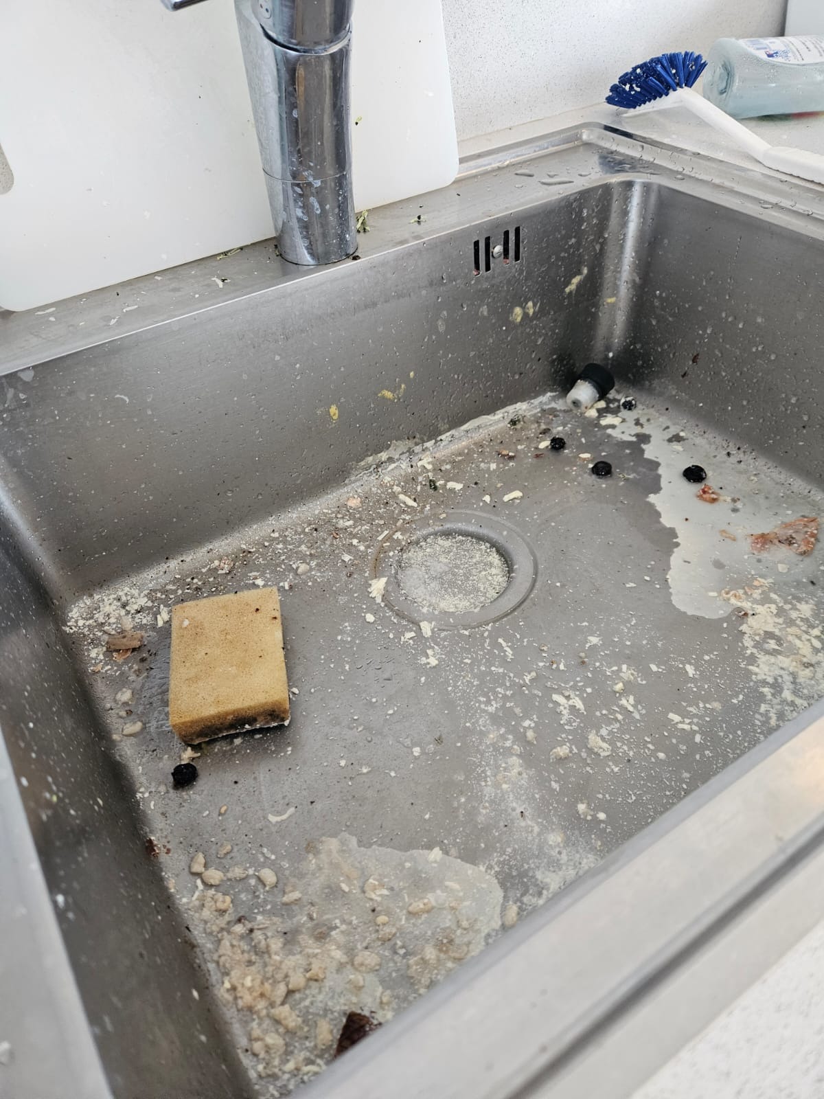 Zanieczyszczony zlew kuchenny ze stali nierdzewnej, widoczna gąbka, resztki jedzenia i zabrudzenia. Potrzeba gruntownego wysprzątania.