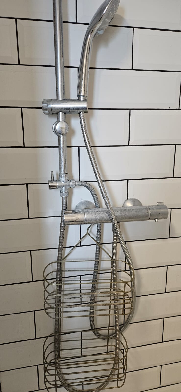 Bateria prysznicowa z półką na kosmetyki na tle białych, prostokątnych płytek z czarną fugą. Widoczne ślady użytkowania na armaturze.