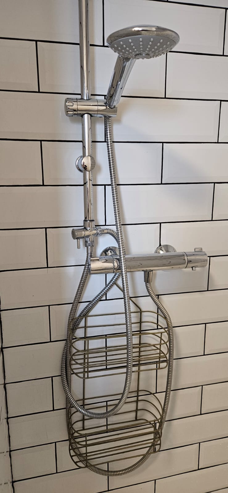 Bateria prysznicowa z deszczownicą i półką na kosmetyki, zamontowana na ścianie z białych, prostokątnych płytek z czarną fugą. Chromowane wykończenie.