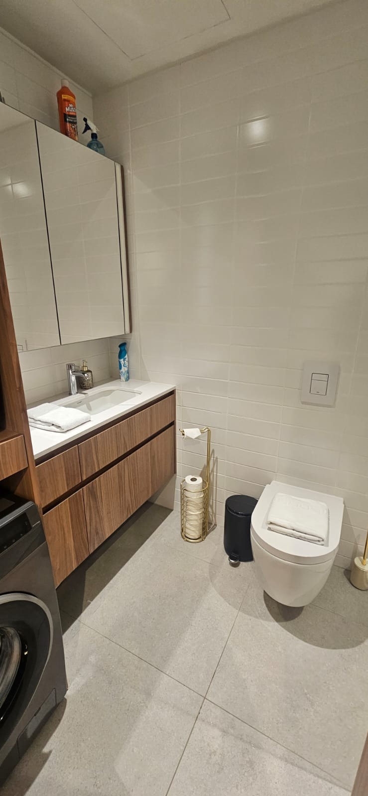 Jasna łazienka po remoncie: białe płytki, drewniana szafka z umywalką, toaleta z deską, złoty stojak na papier toaletowy i szara podłoga. Widok z perspektywy.