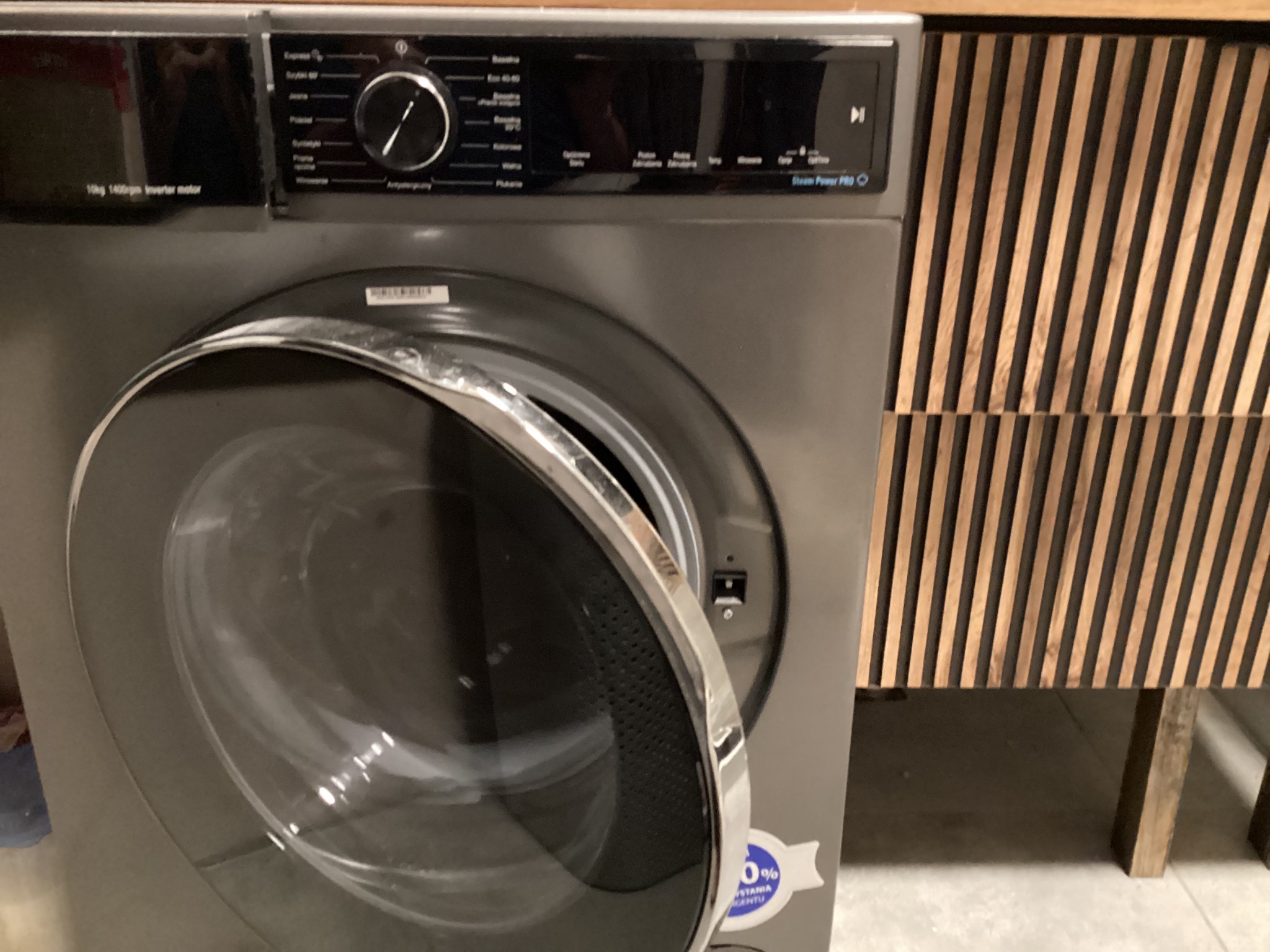 Otwarta pralka automatyczna w odcieniach szarości, z widocznym panelem sterowania i bębnem, stojąca obok szafki z drewnianymi lamelami.