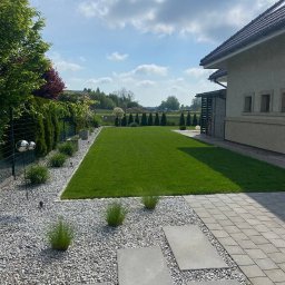 VlaMaks - Zadbany ogród z równo przystrzyżonym trawnikiem, ozdobnymi kamieniami i płytami chodnikowymi. W tle widoczne drzewa i budynek z jasną elewacją.