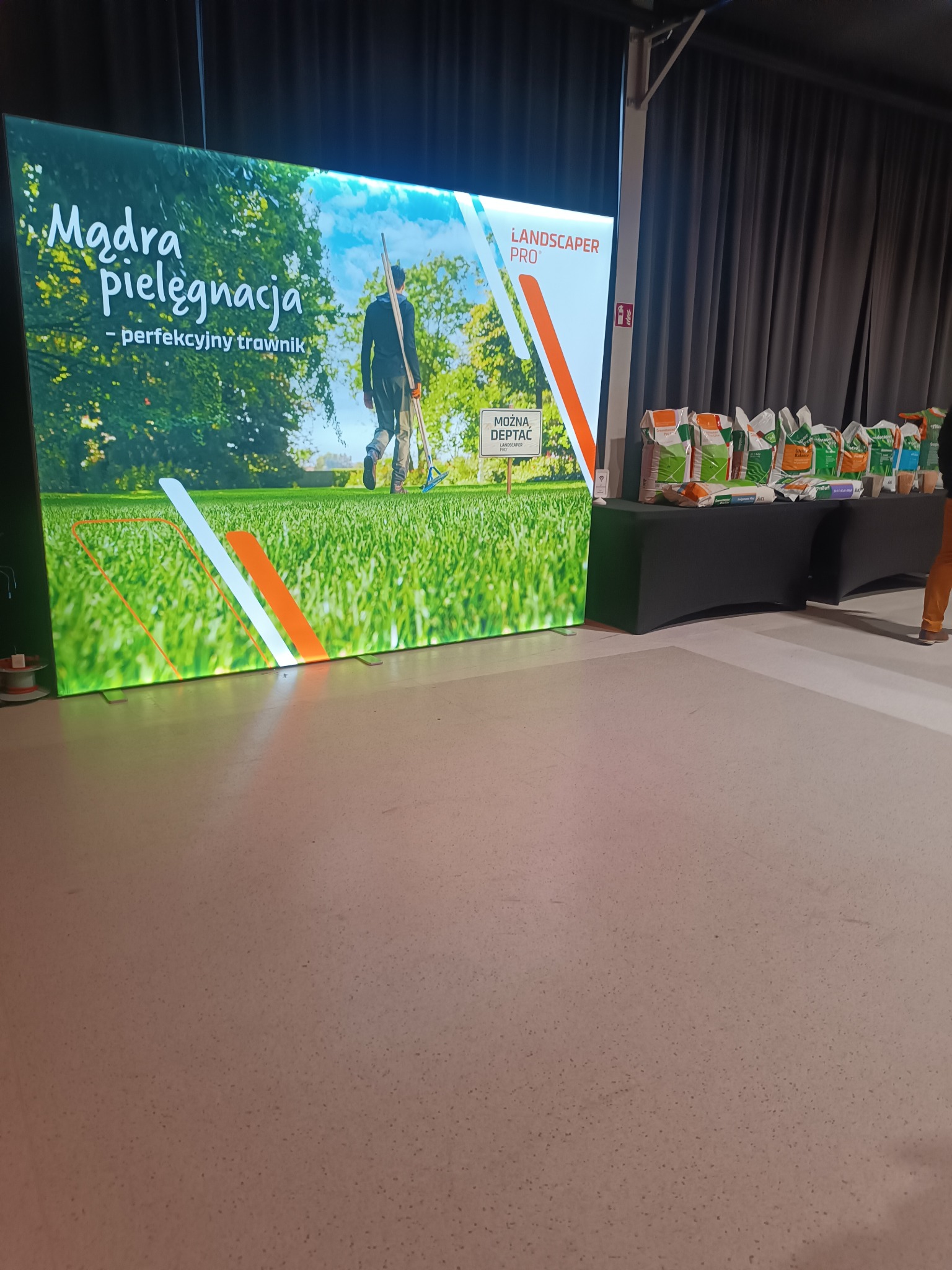Stoisko targowe z podświetlanym banerem 'Mądra pielęgnacja - perfekcyjny trawnik' i produktami do pielęgnacji trawnika na stole. Prezentacja firmy.
