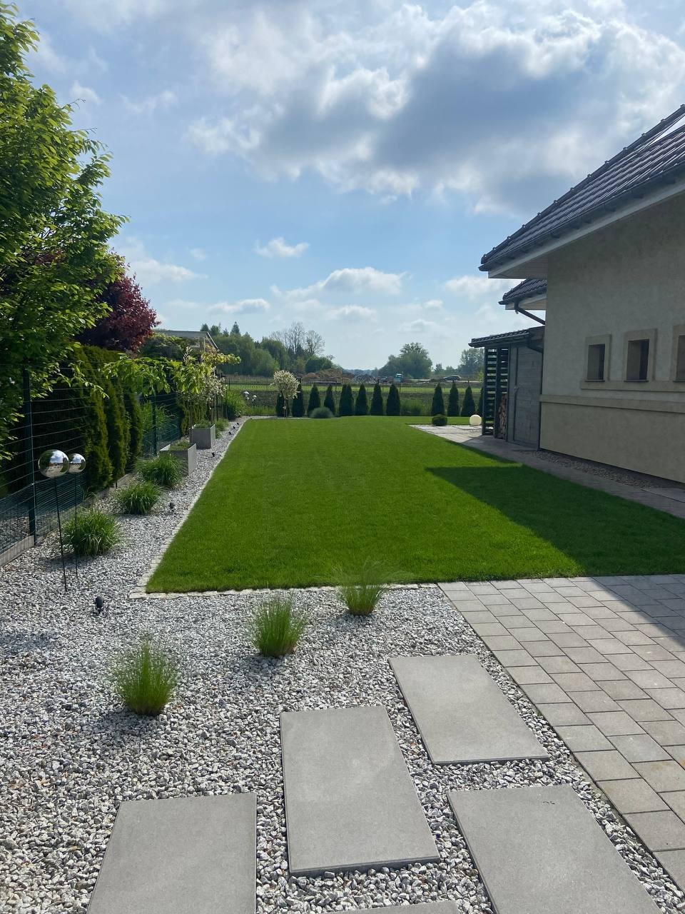 Zadbany ogród z równo przystrzyżonym trawnikiem, ozdobnymi kamieniami i płytami chodnikowymi. W tle widoczne drzewa i budynek z jasną elewacją.