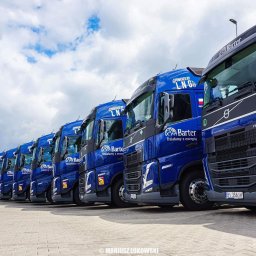 Barter S.A. - Flota niebieskich ciężarówek z logo firmy Barter, z napisem 'Powered by LNG', ustawiona w rzędzie na placu. Widoczny maszt z flagą firmy. Pochmurne niebo w tle.
