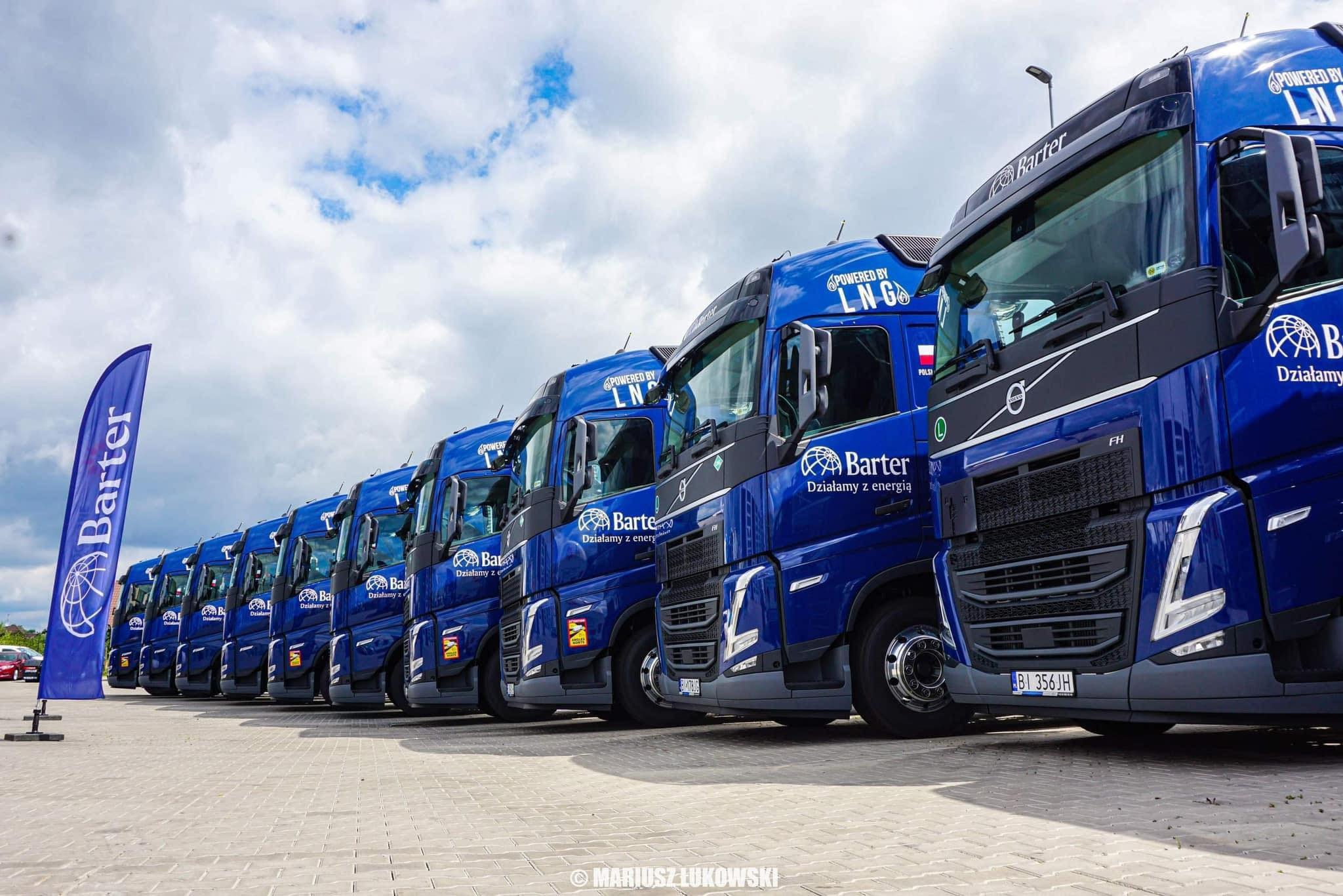 Flota niebieskich ciężarówek z logo firmy Barter, z napisem 'Powered by LNG', ustawiona w rzędzie na placu. Widoczny maszt z flagą firmy. Pochmurne niebo w tle.