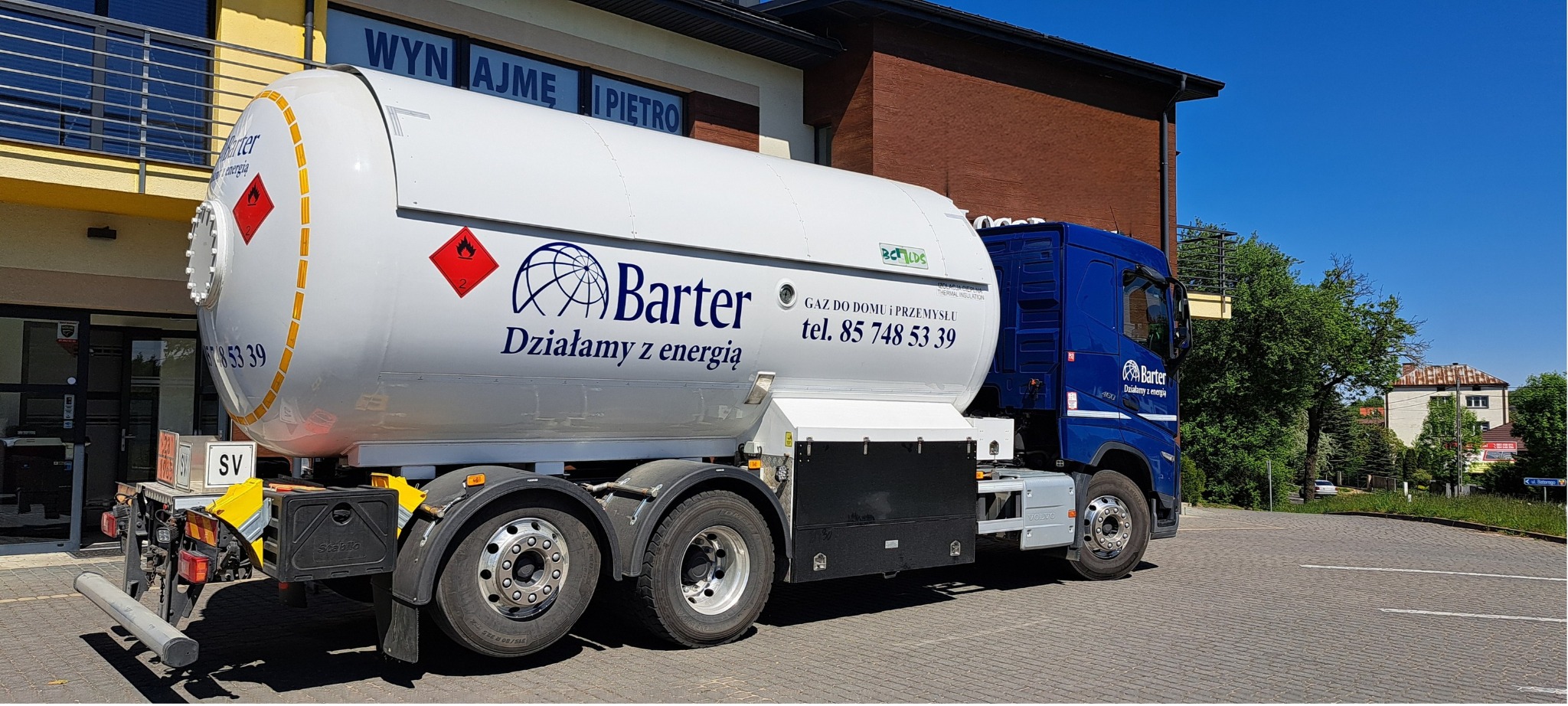 Cysterna z logo firmy Barter, dostarczająca gaz do domów i przemysłu, zaparkowana na brukowanym placu w słoneczny dzień. Widoczny budynek w tle.
