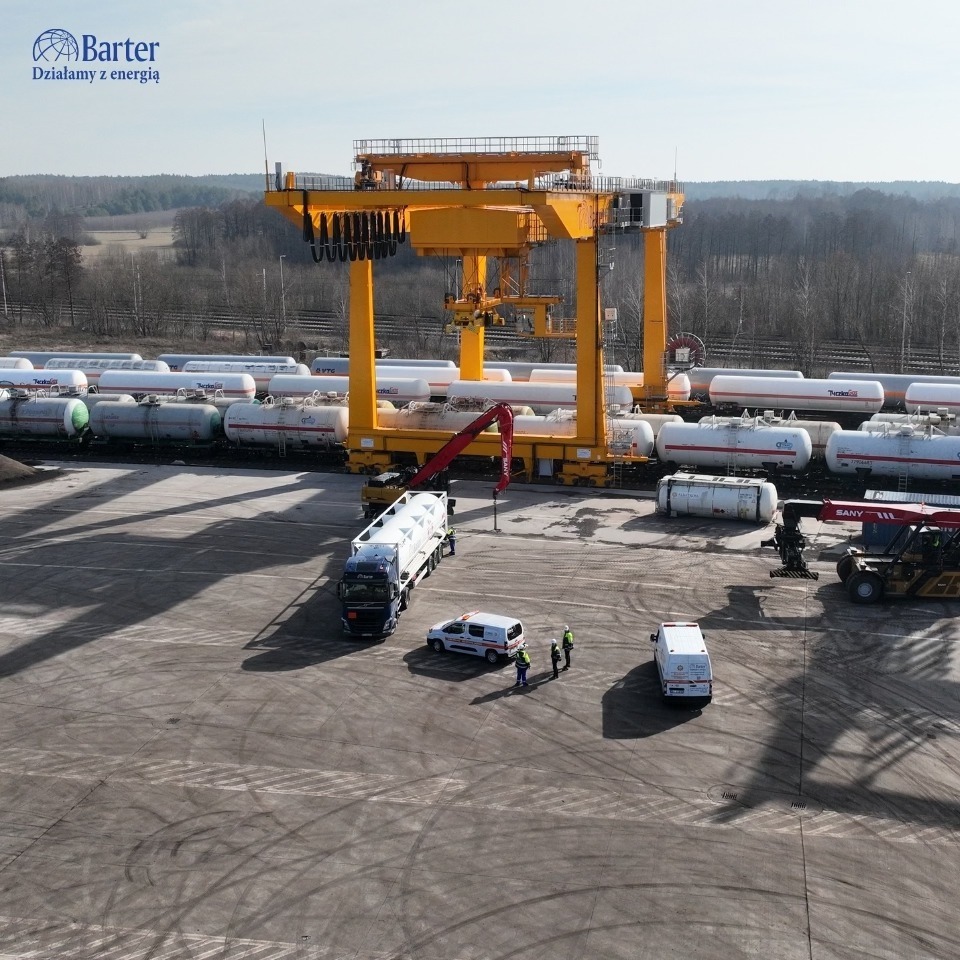 Przeładunek zbiornika z cysterny kolejowej na ciężarówkę za pomocą suwnicy bramowej. Widoczni pracownicy w odblaskowych kamizelkach i pojazdy.