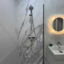 Modena Design - Nowoczesna kabina prysznicowa z marmurowymi płytkami, szklaną ścianką i okrągłym lustrem z podświetleniem LED. Minimalistyczny design, czyste linie, jasne kolory.