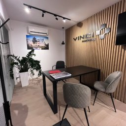 MODENA SPÓŁKA Z OGRANICZONĄ ODPOWIEDZIALNOŚCIĄ - Nowoczesne biuro z drewnianą ścianą, czarnym stołem i szarymi krzesłami. Na ścianie logo firmy i telewizor. Minimalistyczny design z roślinami i oświetleniem punktowym.