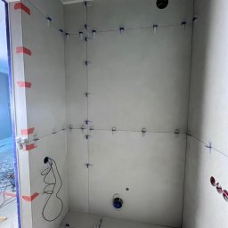 Modena Design - Ściany w trakcie układania płytek w łazience, z użyciem systemu poziomowania. Widoczne przewody elektryczne i otwory na instalacje. Szare płytki, niebieski sufit.