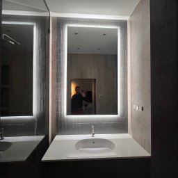 MODENA SPÓŁKA Z OGRANICZONĄ ODPOWIEDZIALNOŚCIĄ - Nowoczesna łazienka z podświetlanym lustrem i umywalką nablatową. Szare płytki i ciemne meble tworzą elegancki kontrast. Widoczne detale wykończenia.