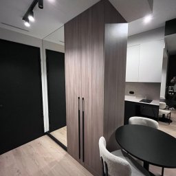 MODENA SPÓŁKA Z OGRANICZONĄ ODPOWIEDZIALNOŚCIĄ - Nowoczesne wykończenie wnętrza: drewniana szafa w zabudowie, czarne drzwi, okrągły stół i odbicie kuchni w lustrze. Minimalistyczny design z akcentami bieli i czerni.