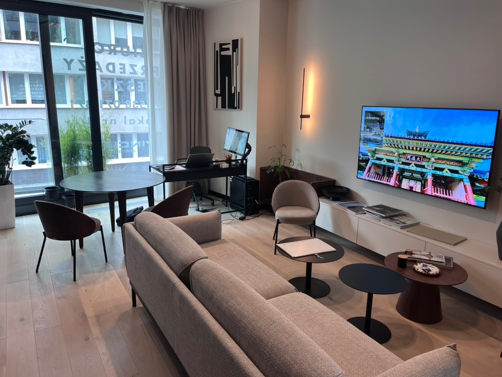 Jasne, przestronne wnętrze salonu z drewnianą podłogą, szarą sofą i telewizorem z widokiem na azjatycką architekturę. Minimalistyczny design, duże okna z widokiem na miasto.