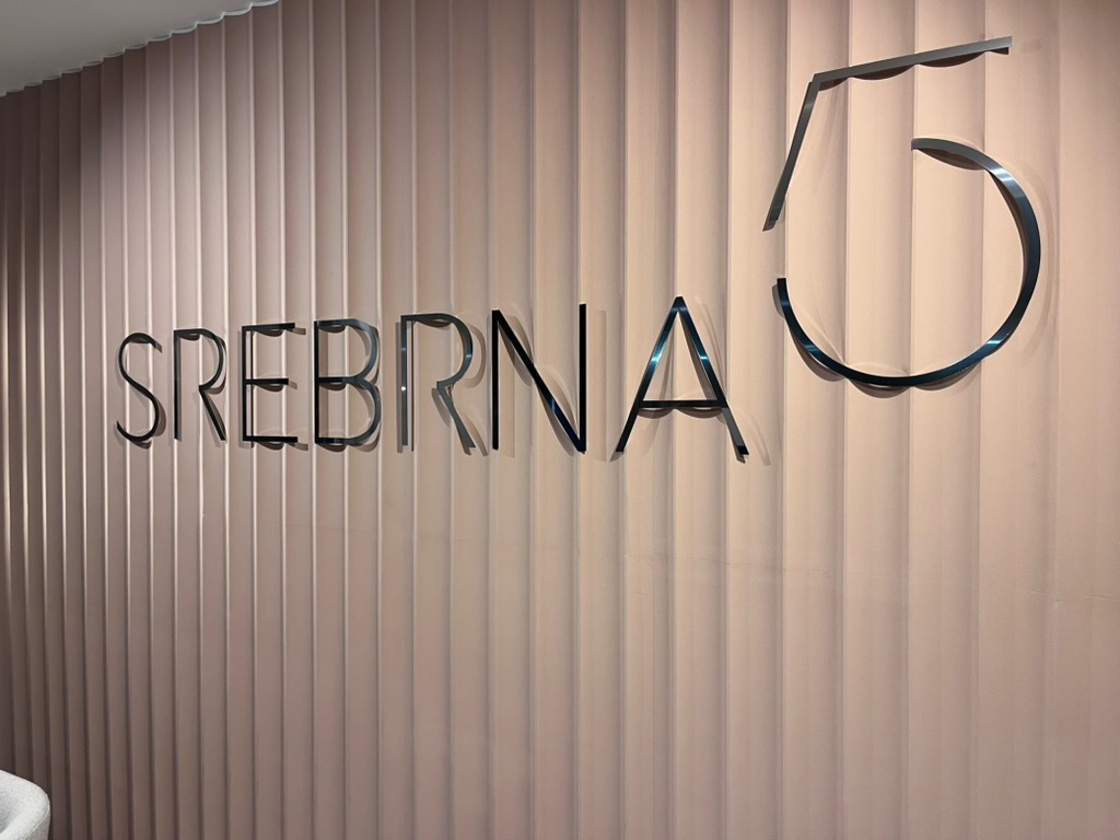 Metalowy napis 'SREBRNA 5' na beżowej, pionowo żłobionej ścianie. Minimalistyczny design, nowoczesny styl, delikatne oświetlenie.