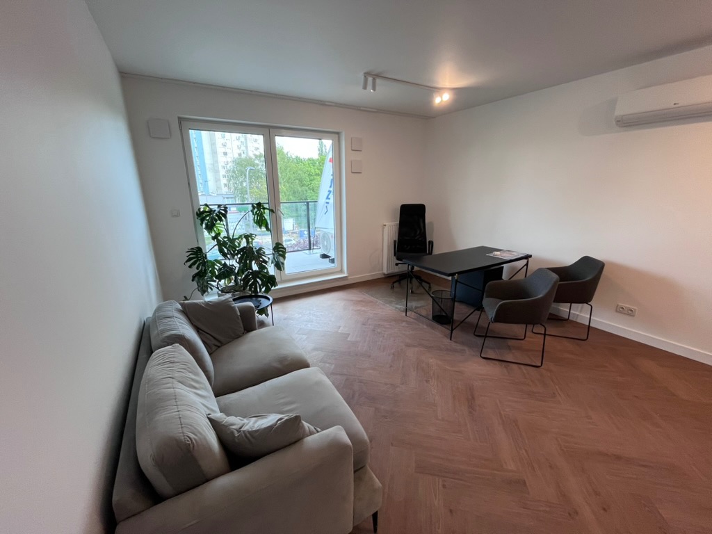 Jasne biuro z parkietem w jodełkę, sofą, biurkiem i krzesłami. Duże okno z widokiem na zieleń. Minimalistyczny design, neutralna kolorystyka, nowoczesne oświetlenie.