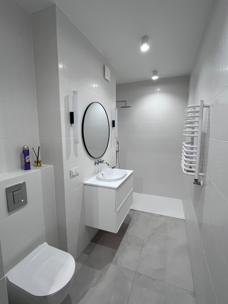 Minimalistyczna łazienka w bieli i szarości: owalne lustro, podwieszana szafka z umywalką, grzejnik drabinkowy i kabina prysznicowa typu walk-in.