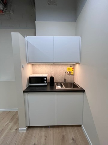 Nowoczesny aneks kuchenny z białymi szafkami, czarnym blatem i oświetleniem LED. Widoczny zlew, bateria, mikrofalówka i ekspres do kawy. Minimalistyczny design.