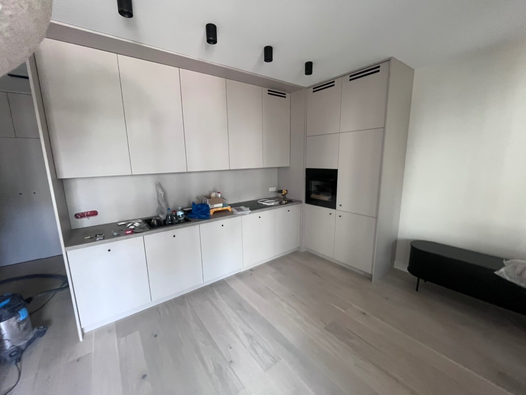 Nowoczesna, jasna kuchnia z zabudową meblową pod sufit i drewnianą podłogą. Widoczny fragment odkurzacza i narzędzia na blacie. Minimalistyczny design.