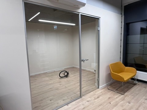 Nowoczesne biuro z przeszkloną ścianą działową i żółtym krzesłem. Minimalistyczny design, jasne drewniane panele podłogowe, widoczne okablowanie.